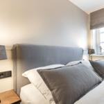 Bedroom with bed, nightstand, and lamp. Window with roller shade and plants.