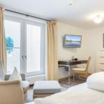 Schlafzimmer mit Bett, Sessel, Schreibtisch und TV. Fenster mit Vorhängen.