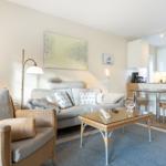 Wohnzimmer mit Sofa, Sessel und Esstisch. Küche mit weißen Schränken.