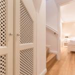 Hallway with wooden stairs and white doors with lattice panels. Bedroom with bed and nightstand in background.