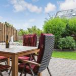 Terrasse mit Holztisch, Stühlen und Weinflasche. Hintergrund: Grünflächen und Holzzaun.