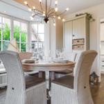 Runder Tisch mit Stühlen, Fenster und Küche im Hintergrund.