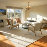 Living room with sofa, chairs, and fireplace. Windows, TV, and chandelier visible.