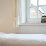 Ein Bett mit weißer Bettwäsche, eine Wandlampe und ein Fenster mit Vorhang und Korb.