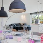Essbereich mit Tisch, Stühlen und großen Fenstern. Küche mit weißen Schränken. Wohnbereich mit Sofa.