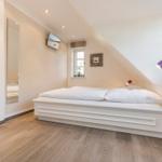 Bedroom with sloped ceiling, white bed, and wooden floor. Windows, mirror, and nightstand with lamp.