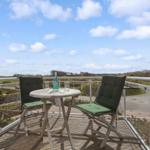 Terrasse mit Tisch und zwei grünen Stühlen. Blick auf Felder und Himmel.