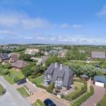 Haus mit Garten und Terrasse, umgeben von Bäumen und weiteren Häusern unter blauem Himmel.