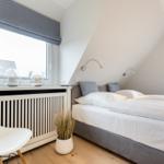 Doppelbett mit weißen Laken, zwei Fenster mit Rollladen und weißer Stuhl.