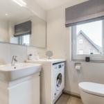 Badezimmer mit Waschmaschine, Spiegel und Toilette. Fenster mit Rollladen.