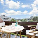 Terrasse mit Tisch und Stühlen, Blick auf Dächer und Grünflächen unter blauem Himmel.