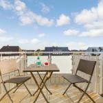 Terrasse mit Tisch und zwei Stühlen. Hintergrund: Dächer und blauer Himmel mit Wolken.