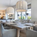 Dining area with wooden table, chairs, and large windows. Pendant lights and kitchen area in background.