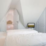 Schlafzimmer mit Dachschrägen, Bett und TV. Tür führt in Bad mit Rundfenster.