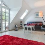 Wohnzimmer mit Sofa, Sessel, Tisch und rotem Teppich. Große Glastür führt auf Balkon.