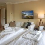 Two beds with towels, sofa, TV, and floor lamp in a bright room.