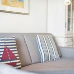 Graße Couch mit Kissen, eines mit Segelbootmotiv, vor weißer Wand und goldener Stehlampe.
