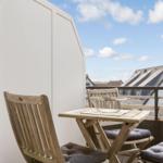 Terrace with wooden table and two chairs with cushions. View of rooftops under blue sky.