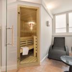 Sauna with wooden bench and glass door. Adjacent area with lounge chairs and table.