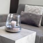 A lit candle in a glass on a table in front of a sofa with cushions.