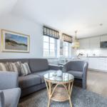 Living room with sofa, armchair, and kitchen area. Wooden floor and large windows.