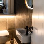 Badezimmer mit Toilette, Waschbecken und Spiegel. Fenster mit Blumen auf Sims.