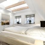 Schlafzimmer mit Dachschrägen, zwei Fenstern und einem Bett mit weißen Kissen.
