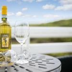 A bottle of white wine and two glasses sit on a terrace table with a view of green hills.