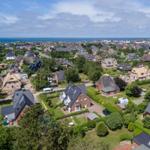 Aerial view of a coastal town with houses, greenery, and the sea in the background.
