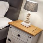 Nachttisch mit Holzoberfläche, Lampe und Bett im Hintergrund.