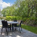 Terrasse mit Tisch und Stühlen neben Haus, Grünfläche und Bäume im Hintergrund.