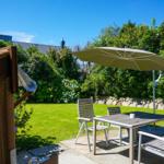 Terrace with table, chairs, and umbrella. Lawn and hedge in the background.