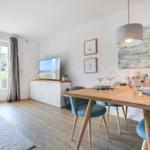 Essbereich mit Holztisch, blauen Stühlen und TV. Fenster zur Außenterrasse.