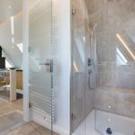 Dusche mit Glaswand und Duschbrause. Im Hintergrund Schlafzimmer mit Fenster.