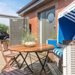 House with terrace, wooden table, chairs, and beach chair. Background: black car and red wall.