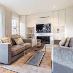 Living room with two sofas, TV, and fireplace. Wooden floor and large windows.