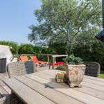 Terrasse mit Tisch, Stühlen und Liegestühlen. Hintergrund: Baum und Hecke.