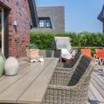 Terrasse mit Tisch, Stühlen und Sonnenschirm. Hintergrund: grüne Hecke und Haus.