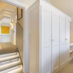 Schlafzimmer mit weißem Kleiderschrank und Nachttisch. Treppenhaus mit beleuchteten Stufen und gelbem Wandbild.