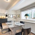 Dining room with table, chairs, and window. Kitchen in background. White walls and wooden floor.