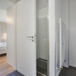 Badezimmer mit Dusche und Heizkörper. Durchgang in Schlafzimmer mit Bett und Fenster.