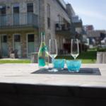 Außenbereich mit Holztisch, Flasche und Gläsern. Hintergrund: Steinhaus mit Balkonen.