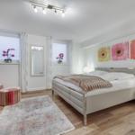 Schlafzimmer mit Bett, zwei Fenstern, Spiegel und Blumenbildern an der Wand.