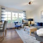 Living and dining area with sofa, coffee table, and TV. Wooden floor and large windows.