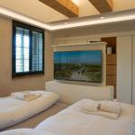 Two beds with white linens, TV, and window with blinds.