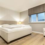 Bedroom with bed, nightstand, lamp, and armchair. Wooden floor and window with roller blind.