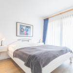 Schlafzimmer mit weißem Bett, grauer Decke und blauen Vorhängen.