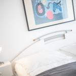 Ein Bett mit weißen Kissen und einer grauen Bettdecke. An der Wand hängt ein abstraktes Bild.