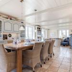 Dining room with wooden table, chairs, and kitchen area. Living area with sofa and fireplace in the background.