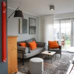 Living room with sofa, armchair, and dining table. Red wall clock and large windows.